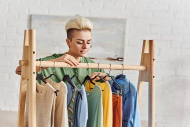 Genç ve mutlu bir kadın oturma odasındaki raflarda duran şık günlük kıyafetlere bakıyor, ikinci el, bilinçli yaşam tarzı, sürdürülebilir moda ve dikkatli tüketim konsepti