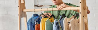 Kısmen gülümseyen ve dövmeli bir kadının evde askıların üzerinde şık gündelik kıyafetlerle durması, sürdürülebilir moda ve dikkatli tüketim konsepti, afiş