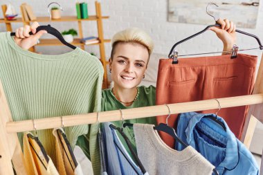 thrift store finds, overjoyed woman with trendy hairstyle showing stylish jumper and pants while looking at camera  near rack with clothes, sustainable fashion and mindful consumerism concept clipart