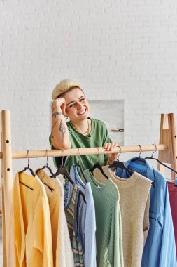 Tasasız dövmeli, modaya uygun saç stili olan bir kadın kameraya gülümserken askılarda modaya uygun günlük giysiler, sürdürülebilir moda ve dikkatli tüketim konseptiyle ayakta duruyor.