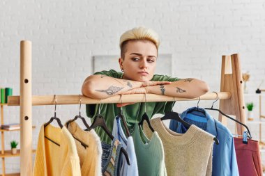 Düşünceli ve üzgün dövmeli bir kadın, modern oturma odasında, moda ve düşünceli tüketim konseptindeki askılarla, modaya uygun kıyafetlerle raflarda öğreniyor.