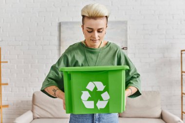 smiling tattooed woman with trendy hairstyle looking in green plastic box with recycling sign while standing at home, reduce waste, sustainable living and environmentally friendly habits concept clipart