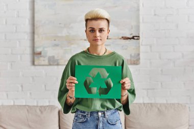 young woman in stylish casual clothes, with trendy hairstyle holding green paper card with cutout recycling symbol and looking at camera, sustainable and environmentally conscious lifestyle concept clipart