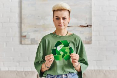 positive tattooed woman with trendy hairstyle showing green recycling sign with globe while smiling at camera in modern living room, sustainable living and environmental awareness concept clipart