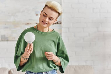 cheerful young woman in casual clothes looking at energy saving light bulb at home, trendy hairstyle, tattoo, positive emotion, sustainable lifestyle and environmentally conscious concept clipart