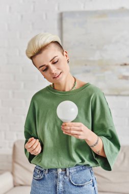 green living, young tattooed woman with trendy hairstyle and happy face holding energy saving light bulb at home, sustainable lifestyle and environmentally conscious concept clipart