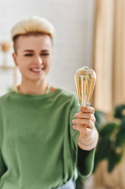 cheerful woman with trendy hairstyle, in casual clothes smiling and standing with energy saving light bulb at home on blurred background, sustainable lifestyle and environmentally conscious concept clipart