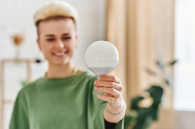 energy saving light bulb in hand of overjoyed young woman with trendy hairstyle standing at home on blurred background, sustainable lifestyle and environmentally conscious concept clipart