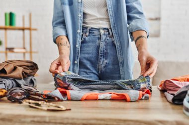 partial view of young and tattooed woman in casual clothes sorting wardrobe items on table in living room, exchange market, eco-friendly swaps, sustainable living and mindful consumerism concept clipart