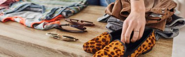 partial view of woman placing fashionable animal print shoes near sunglasses and pre-loved clothes on table at home, eco-friendly swaps, sustainable living and mindful consumerism concept, banner clipart