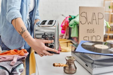 partial view of young and tattooed woman holding electric toaster near vinyl record player, cezve and second-hand clothes during swap not shop event, sustainable living and circular economy concept clipart
