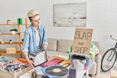 young tattooed woman with electric toaster near vinyl record player, second-hand clothes and carton card with swap not shop lettering at home, sustainable living and circular economy concept clipart