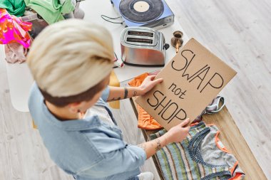 top view of young tattooed woman holding card with swap not shop lettering near clothes, electric toaster, vinyl record player and cezve, sustainable living and circular economy concept clipart