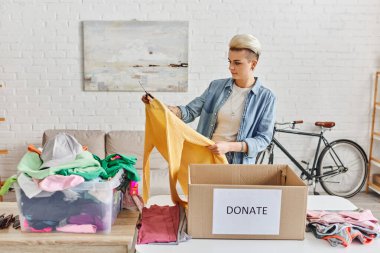 casually styled and tattooed woman holding yellow jumper while sorting clothes near plastic container and carton box with donate lettering, sustainable living and social responsibility concept clipart