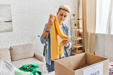 volunteering and charity, trendy and tattooed woman standing with yellow jumper near carton box while sorting clothing for donation at home, sustainable living and social responsibility concept clipart