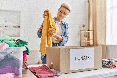 Genç ve şık bir kadın, sarı kazakla plastik konteynırın yanında duruyor, oturma odasında giysi ve bağış kutusu var, sürdürülebilir yaşam ve sosyal sorumluluk kavramı.