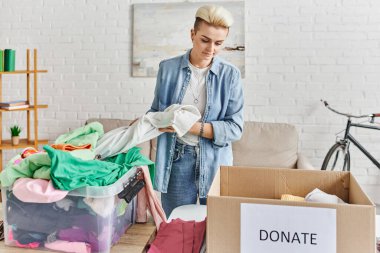 tattooed woman with trendy hairstyle looking at carton box with donate lettering while sorting wardrobe items in modern living room, sustainable living and social responsibility concept clipart