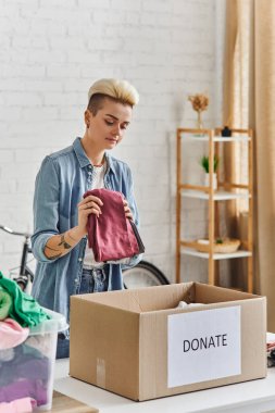donating for a cause, tattooed woman with trendy hairstyle holding clothes near carton box with donate lettering in modern living room, sustainable living and social responsibility concept clipart