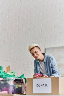 young and joyful woman with trendy hairstyle holding pre-loved clothes and looking at camera near plastic container and donation box at home, sustainable living and social responsibility concept clipart