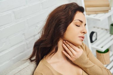 High angle view of young brunette woman in casual jumper massaging lymphatic nodes on neck while sitting on couch at home, self-care ritual and holistic wellness practices concept, tension relief clipart