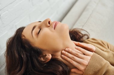 Close up view of relaxed brunette woman in brown jumper massaging neck during lymphatic drainage support and sitting on couch at home, self-care ritual and holistic healing concept, tension relief clipart