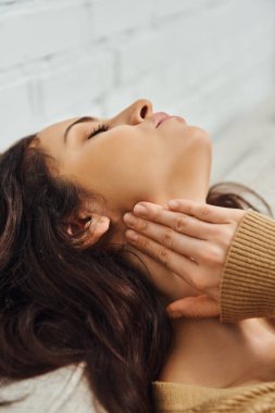 Young brunette woman in brown jumper touching lymphatic node on neck and relaxing during self-massage on couch at home, self-care ritual and holistic healing concept, tension relief clipart