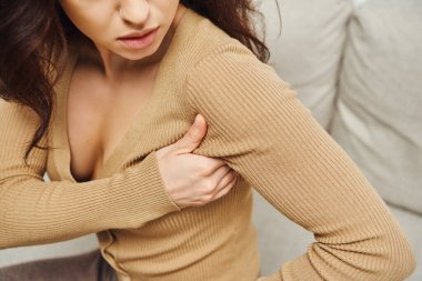 Cropped view of young brunette woman in brown jumper touching lymphatic nodes on armpit for drainage and support on couch at home, enhancing self-awareness and body relaxation concept clipart