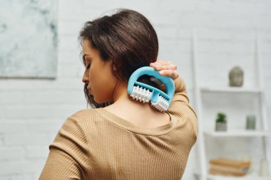Side view of young brunette woman in brown jumper massaging neck and back with handled massager in blurred living room at home, self-care ritual and holistic wellness and health concept clipart