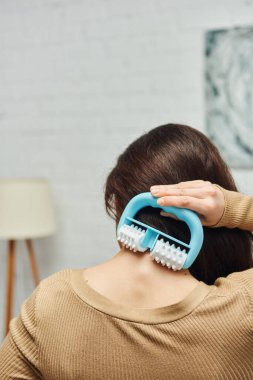 Sırtında kahverengi kazak tutan, masaj yapan ve bulanık oturma odasında kendine masaj yapan genç bir kadın, lenf sistemi desteği ve ev tabanlı masaj, gerilim giderici.