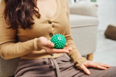 tension relief, cropped view of blurred brunette woman in casual clothes holding manual massage ball while sitting near couch at home, lymphatic system support and home-based massage  clipart