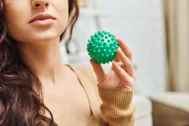Cropped view of young brunette woman in jumper holding manual massage ball while standing in blurred living room at home, lymphatic system support and home-based massage, tension relief clipart