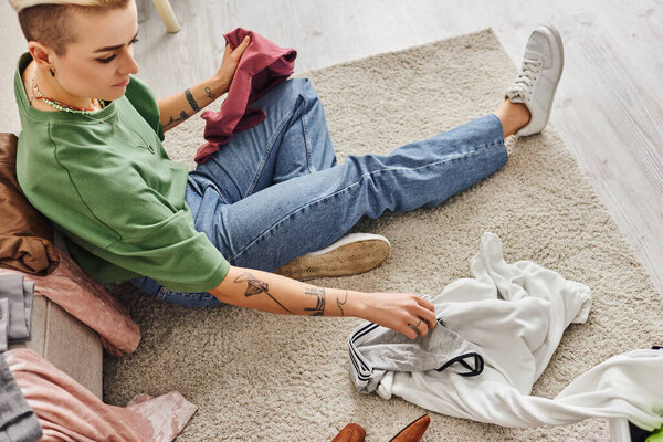 high angle view of young tattooed woman in casual clothes sorting clothes and thrift store finds while sitting on floor carpet in living room, sustainable living and mindful consumerism concept