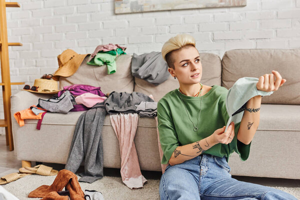 stylish tattooed woman holding cap and looking away while sitting on floor near couch with clothes, wardrobe items sorting, sustainable living and mindful consumerism concept