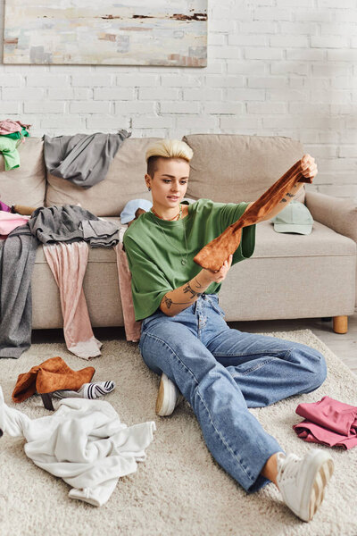 tattooed woman in casual style clothes holding suede boot while sorting belongings and thrift store finds near couch on floor in living room, sustainable living and mindful consumerism concept