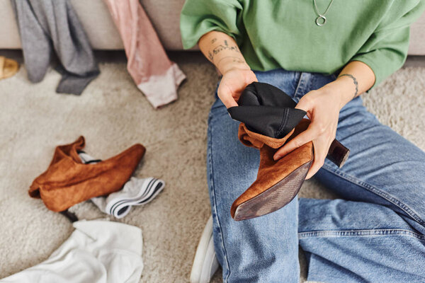partial view of young woman with tattoo sitting on floor in living room and holding suede boot while decluttering clothes at home, top view, sustainable living and mindful consumerism concept