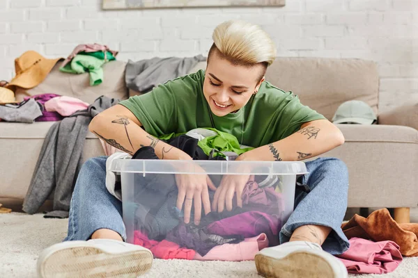 happy tattooed woman sitting on floor and sorting pre-loved items in plastic container near couch at home, trendy hairstyle, clothes decluttering, sustainable living and mindful consumerism concept