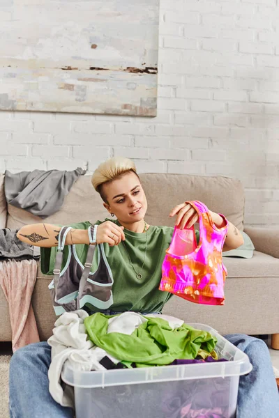 stock image casually styled and tattooed woman looking at colorful tops in modern living room near couch, sorting clothes, reducing wardrobe items, sustainable living and mindful consumerism concept