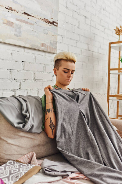 thoughtful young woman with trendy hairstyle and tattoo sitting on couch near wardrobe items and holding grey pants, clothes sorting, decluttering, sustainable living and mindful consumerism concept