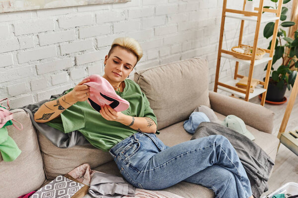 young woman in casual clothes sitting on couch and holding cap near rack and green plant at home, trendy hairstyle, tattoo, wardrobe decluttering, sustainable living and mindful consumerism concept