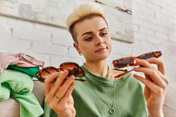 young and pleased woman with trendy hairstyle and tattoo looking at fashionable sunglasses near clothing on couch at home, sorting pre-loved items, sustainable living and mindful consumerism concept