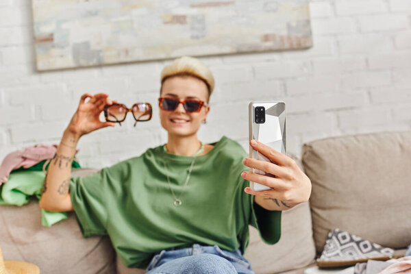 pleased tattooed woman sitting on couch near wardrobe items and taking selfie with sunglasses on smartphone for online exchange, sustainable living and mindful consumerism concept