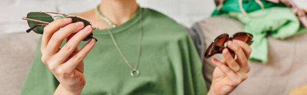 partial view of young woman holding pair of fashionable sunglasses near clothing on blurred background, wardrobe items decluttering, sustainable living and conscious lifestyle concept, banner