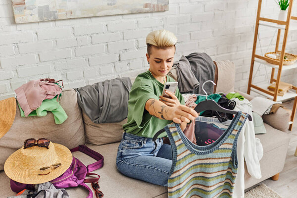 clothing swapping, tattooed woman taking photo of knitted tank top on smartphone on couch at home near straw hats, sunglasses and pre-loved items, sustainable living and mindful consumerism concept 