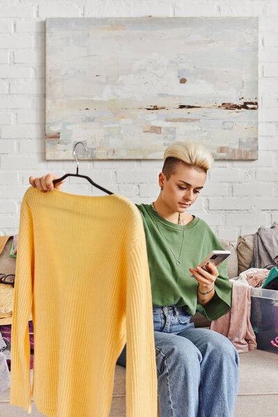 young tattooed woman holding yellow jumper and looking at mobile phone while sitting on couch near wardrobe items, sorting clothes, online swap, sustainable living and mindful consumerism concept