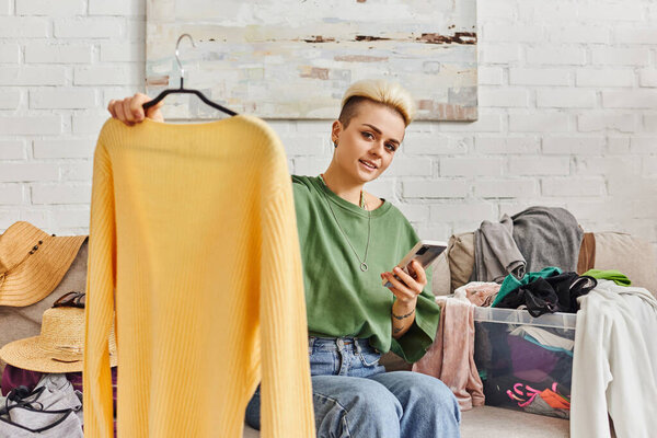 online exchange, tattooed woman smiling at camera, holding smartphone and hanger with yellow jumper, sitting on couch near clothes, sustainable living and mindful consumerism concept