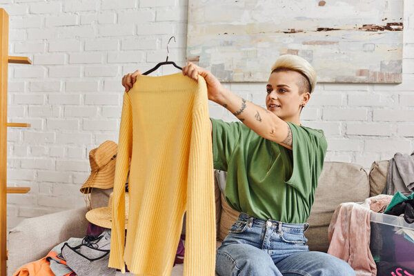 young tattooed woman smiling and looking at yellow trendy jumper while sitting on couch near thrift store finds in modern living room at home, sustainable living and mindful consumerism concept