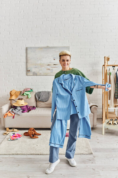 wardrobe and thrift store finds sorting, joyful tattooed woman showing blue pajamas and standing in modern living near couch and rack with clothes, sustainable living and mindful consumerism concept