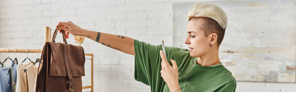 tattooed woman with trendy hairstyle and smartphone taking photo of leather bag near rack with casual style clothes, online swap, sustainable living and mindful consumerism concept, banner