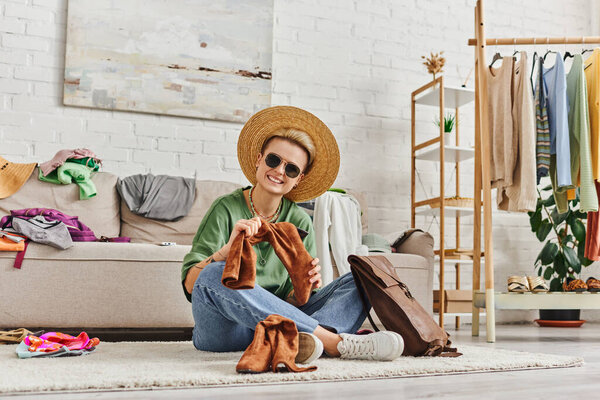 clothes sorting, overjoyed tattooed woman in straw hat and sunglasses looking at camera near suede boots and leather bag on floor at home, sustainable fashion and mindful consumerism concept