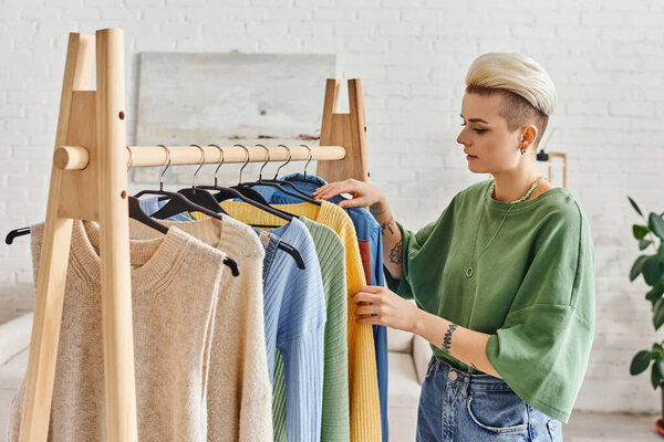 stylish tattooed woman with trendy hairstyle standing near rack with hangers and sorting clothing in modern living room at home, sustainable fashion and mindful consumerism concept
